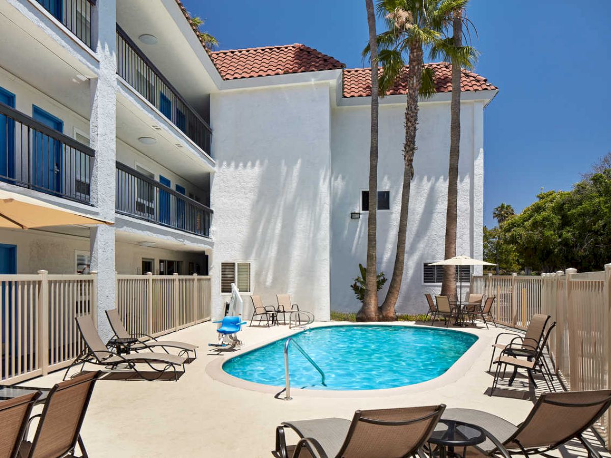 The image shows a small outdoor swimming pool surrounded by lounge chairs and umbrellas, located next to a multi-story white building with balconies.