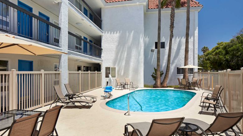 The image shows a small outdoor swimming pool surrounded by lounge chairs and umbrellas, located next to a multi-story white building with balconies.