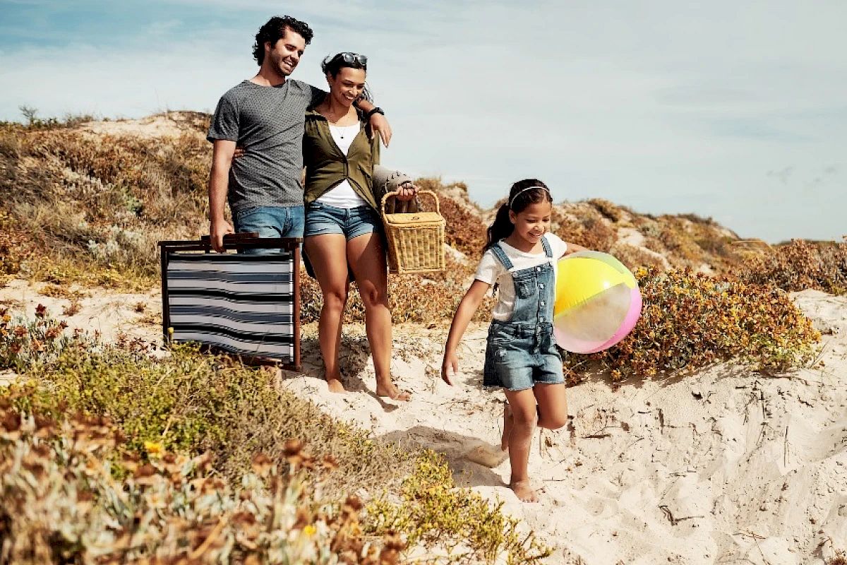 A smiling family with beach accessories is walking through sand dunes toward the beach, enjoying a sunny day.