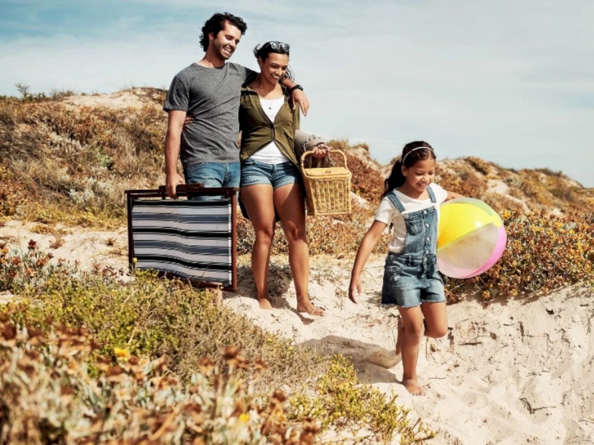 A smiling family with beach accessories is walking through sand dunes toward the beach, enjoying a sunny day.