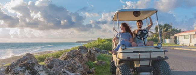 Two people are riding a golf cart along a scenic coastal road with a beautiful sea view and cloudy sky.