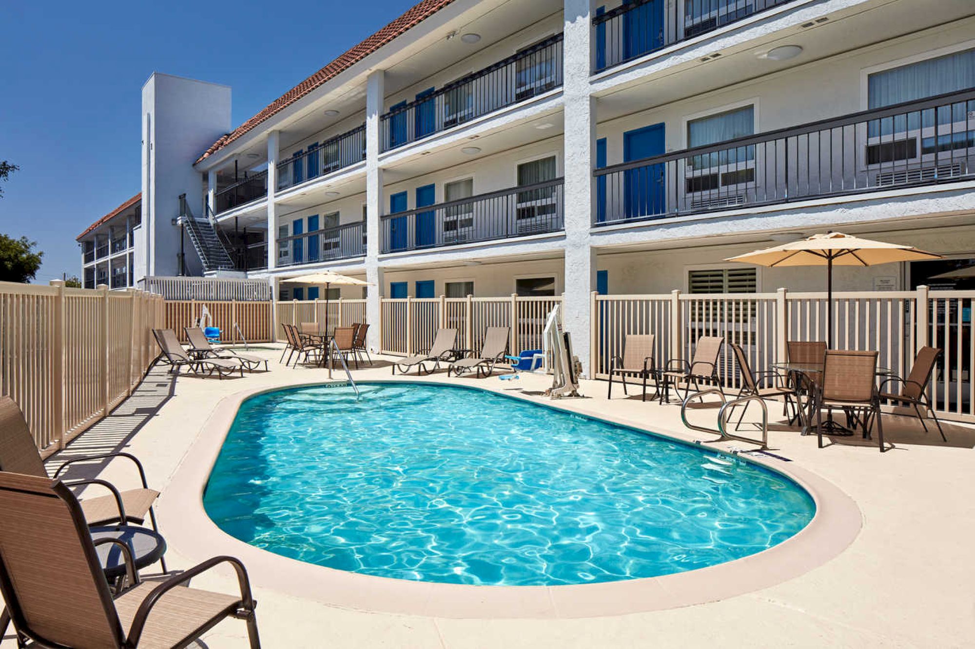 The image shows a swimming pool area with lounge chairs and umbrellas, enclosed by a fence. The setting is adjacent to a multi-story motel.