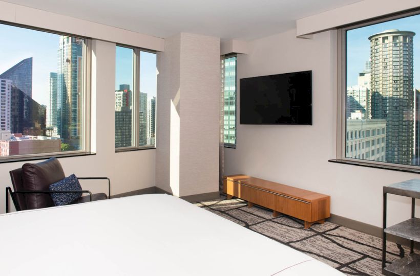 The image depicts a modern hotel room with large windows showcasing a city skyline view. There is a bed, a chair, a TV, and a side table.