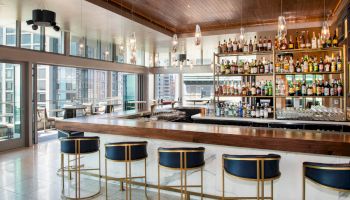 A modern bar with a wooden bar top, blue and gold bar stools, and large windows offering a city view. The bar is stocked with various bottles.