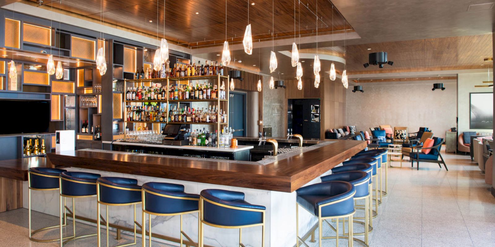This image features a modern bar with a wooden counter, blue bar stools, hanging lights, a well-stocked liquor shelf, and a sleek, open seating area.