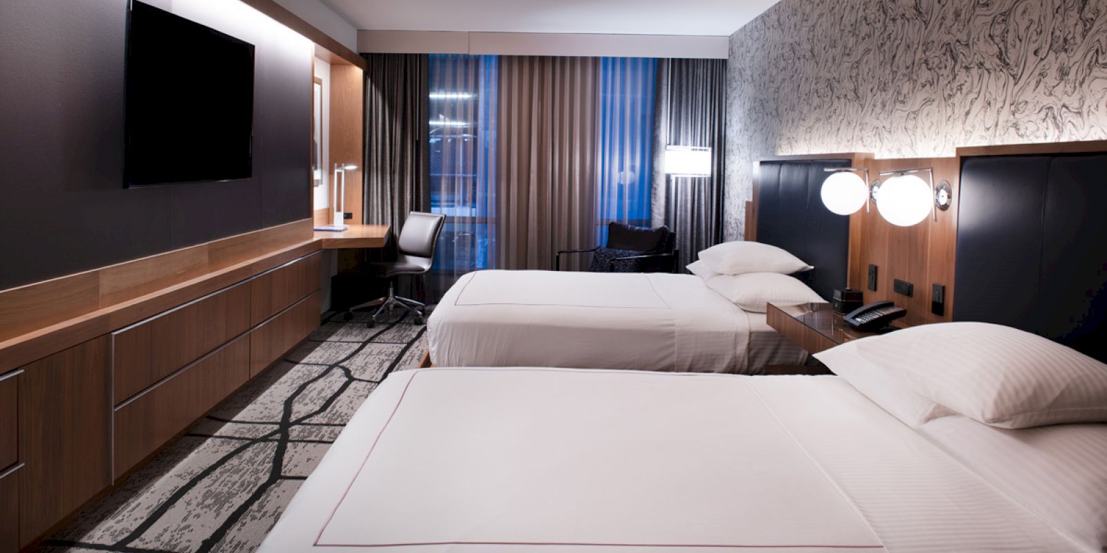 A modern hotel room with two neatly made beds, a flat-screen TV, a desk with a chair, lamps, and large windows with curtains.