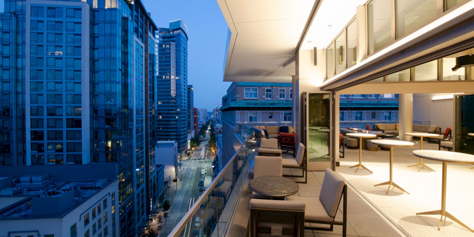A rooftop terrace with seating overlooks a city street with modern high-rise buildings during twilight, creating a tranquil urban setting.