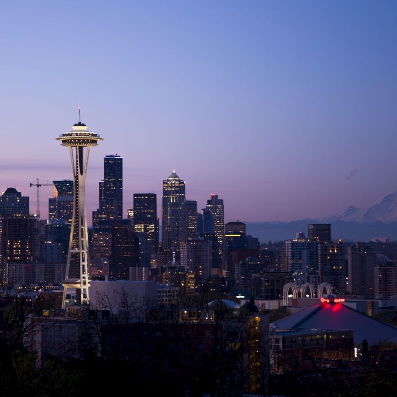 The image shows the Seattle skyline at twilight, featuring the Space Needle and Mount Rainier in the background, with city lights beginning to illuminate.
