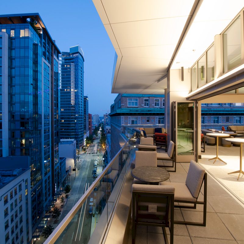 A modern cityscape at dusk with high-rise buildings and a spacious balcony featuring tables and chairs illuminated by warm lighting.