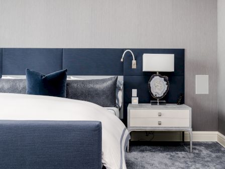 A modern bedroom with a blue and white color scheme, featuring a blue upholstered headboard, a white bed, and a white nightstand with a lamp and decor.