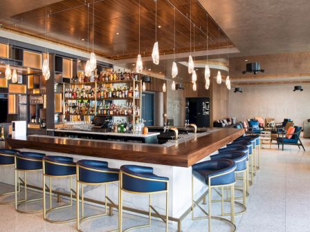 A modern bar with stylish stool seating, a well-stocked bar shelf, hanging lights, and a cozy lounge area in the back, featuring contemporary decor.