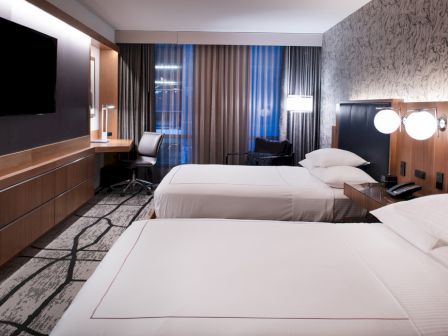 The image shows a modern hotel room with two neatly made beds, a flat-screen TV, a desk, and a chair by a window with curtains.