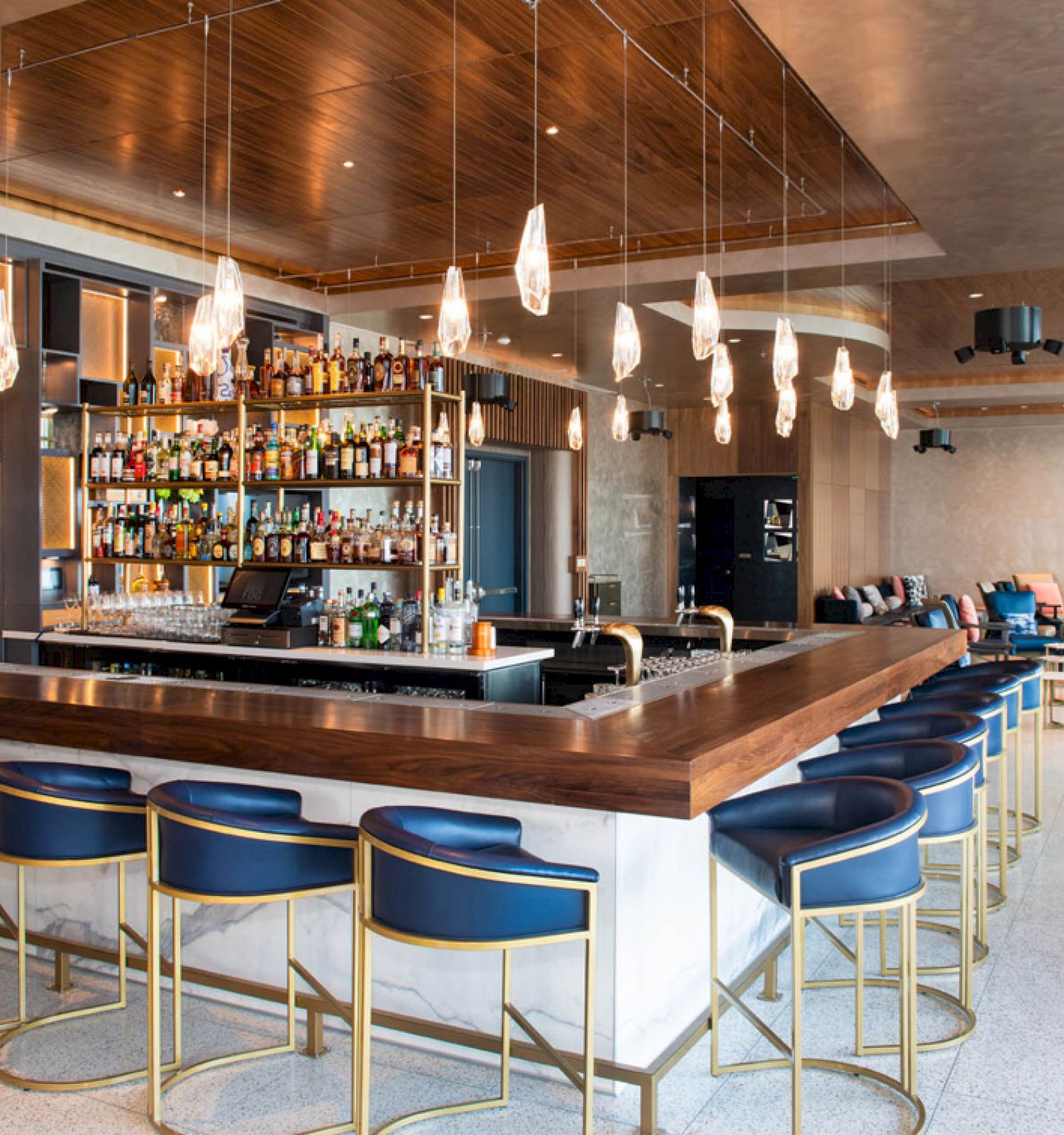 A chic bar area with a modern design, featuring a wooden counter, blue and gold bar stools, pendant lights, and a fully stocked bar.