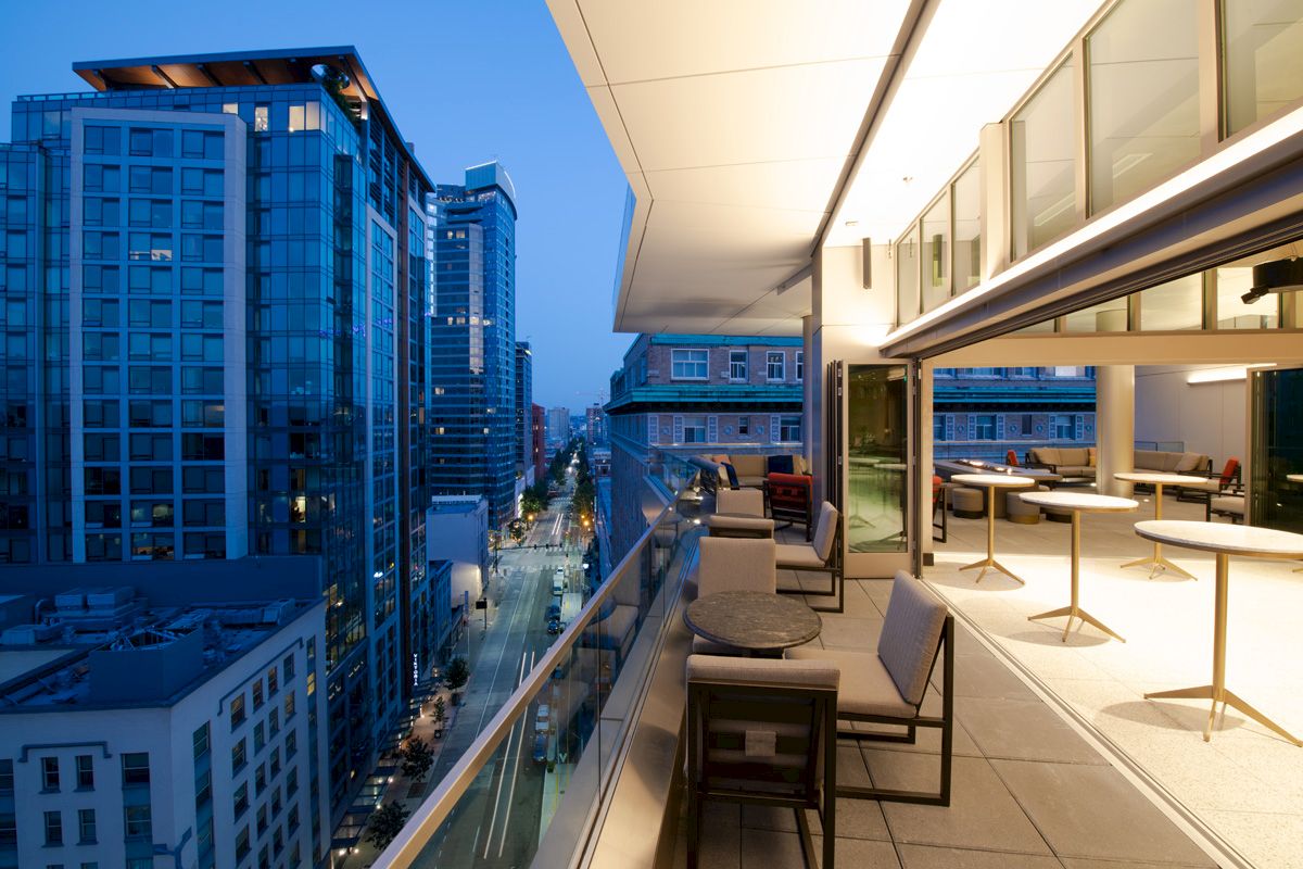 A modern balcony overlooks a city street lined with tall buildings, showcasing a twilight view. The balcony has outdoor seating and tables.