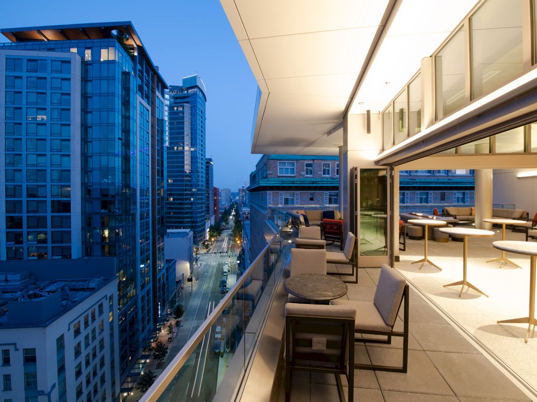 A modern balcony overlooks a city street lined with tall buildings, showcasing a twilight view. The balcony has outdoor seating and tables.