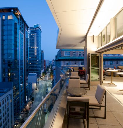 This image shows a modern cityscape viewed from a rooftop terrace at dusk, with tables and chairs on the patio and high-rise buildings in the distance.