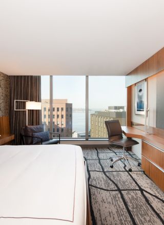 A modern hotel room with a bed, large TV, desk with chair, and a seating area. The window offers a view of buildings and possibly a waterfront.