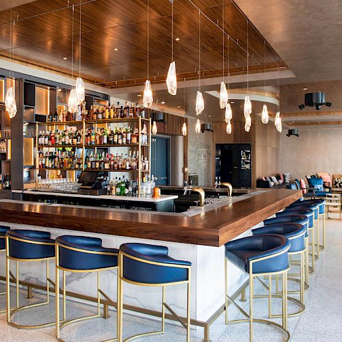 A modern bar with stylish seating, a well-stocked liquor shelf, pendant lights, a large counter, and a cozy lounge area in the background.