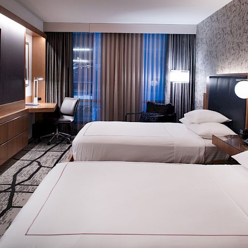 The image shows a modern hotel room with two neatly made twin beds, a flat-screen TV, a desk, and large windows with curtains.