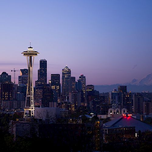 A cityscape at dusk featuring the Space Needle, a recognizable landmark, with skyscrapers and Mount Rainier in the background, lit up by evening lights.