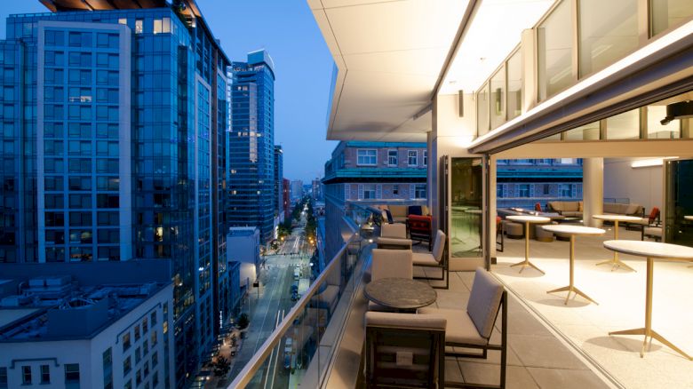 A modern cityscape at dusk with high-rise buildings on the left and a spacious, furnished balcony on the right, illuminated by soft lighting.