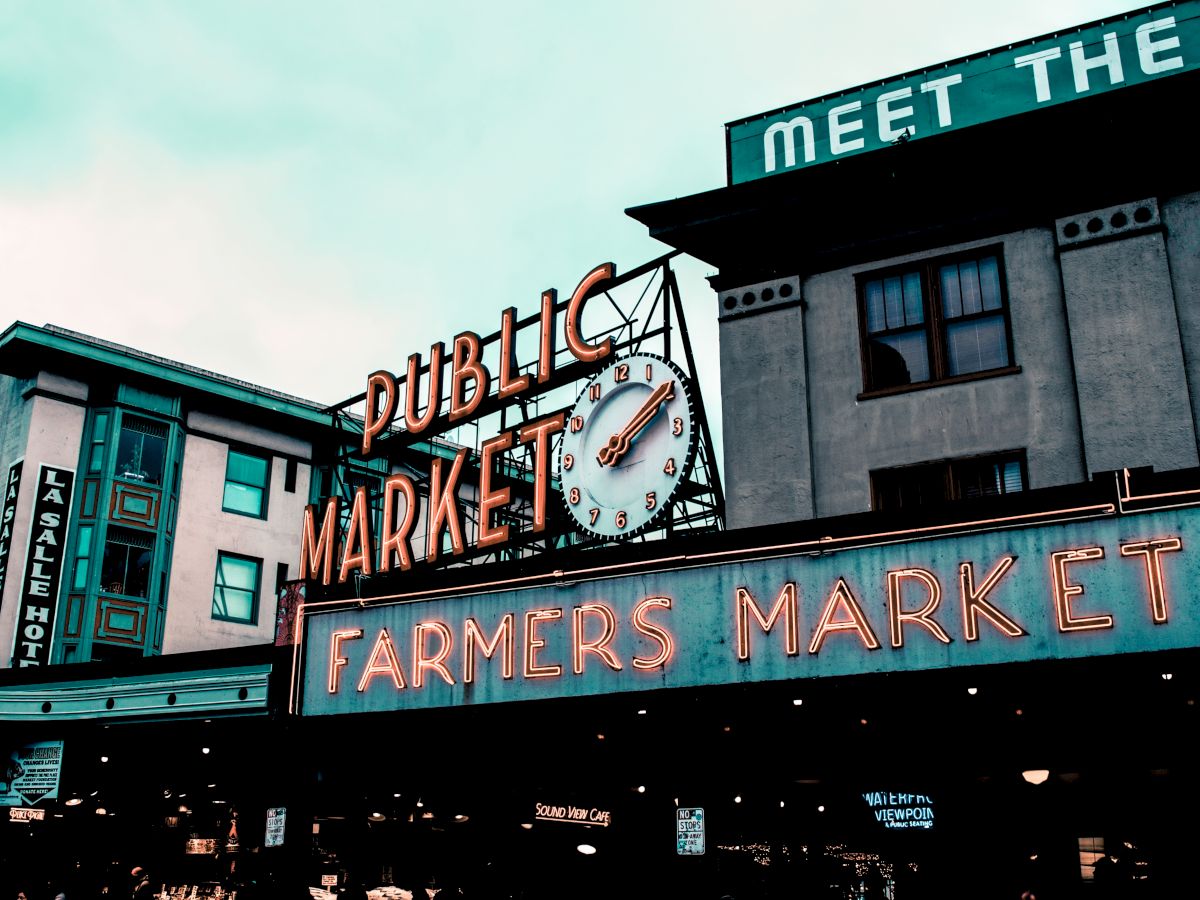 The image shows a public market with neon signs reading "Public Market" and "Farmers Market" on a cloudy day, with vintage architectural buildings in the background.