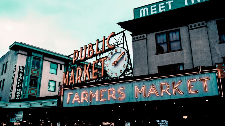 The image shows a public market with neon signs reading "Public Market" and "Farmers Market" on a cloudy day, with vintage architectural buildings in the background.
