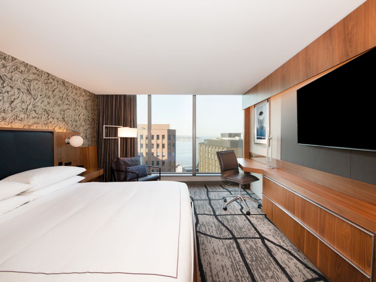 A modern hotel room featuring a large bed, a desk with a chair, a flat-screen TV, a comfortable seating area, and a window with a city view.