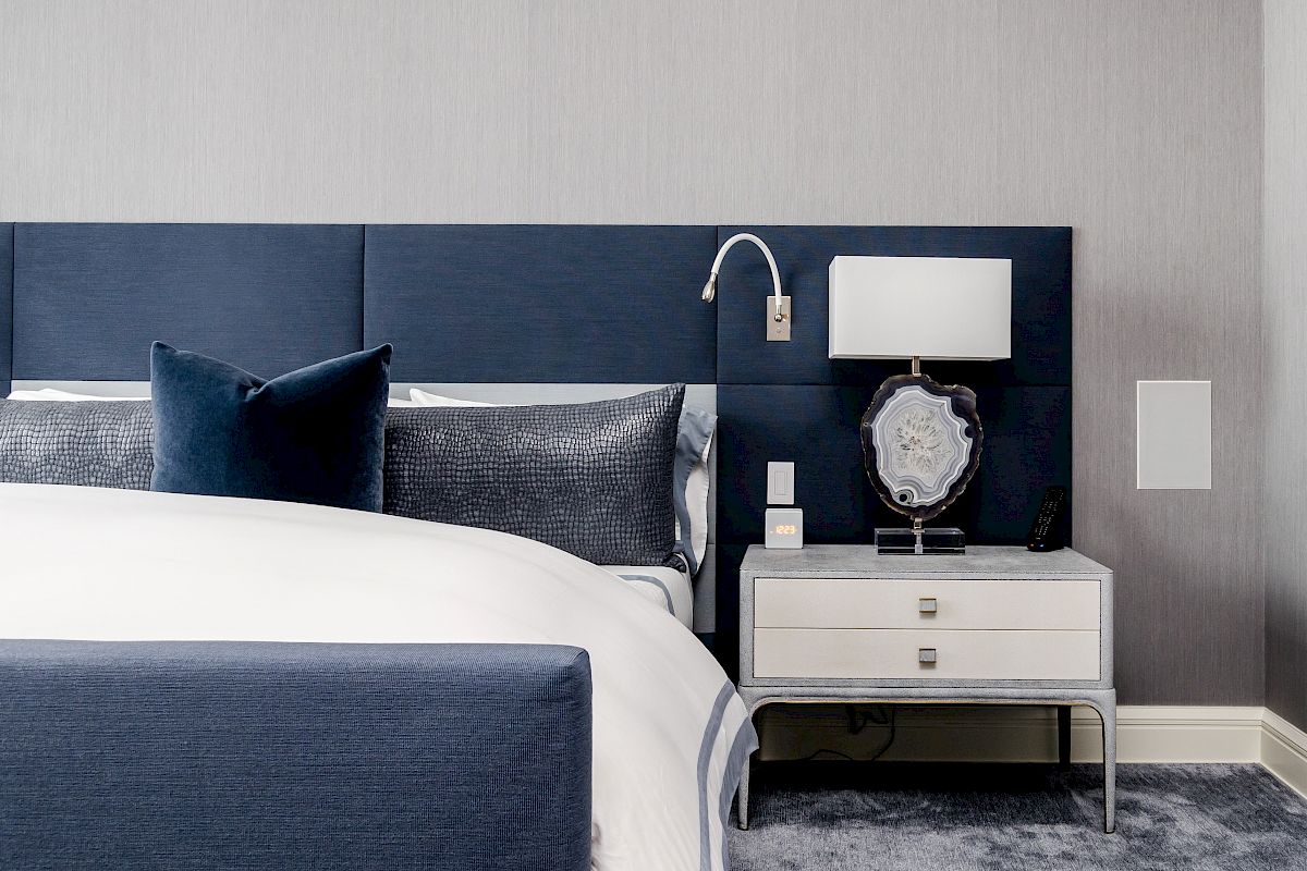 A modern bedroom with a blue upholstered bed, white bedding, a nightstand with a lamp, and a decorative item.