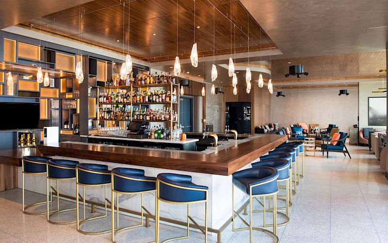 This image shows a modern bar with blue bar stools, a fully stocked backlit liquor shelf, hanging lights, and a spacious seating area in the background.