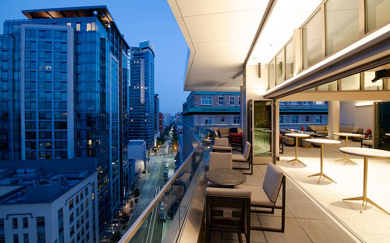 This image shows a modern urban balcony at dusk with seating and a view of tall buildings and a city street below.