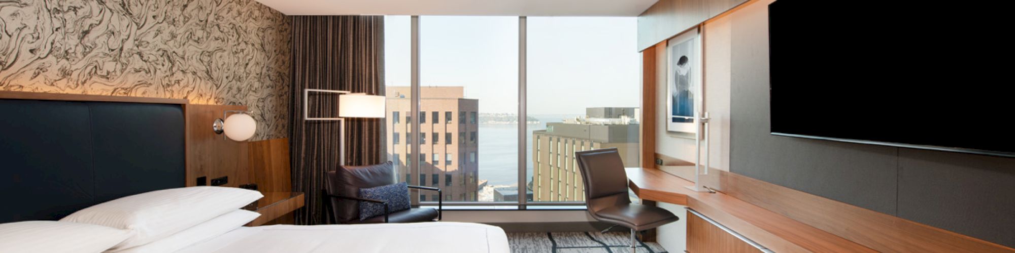 The image shows a modern hotel room with a large bed, a flat-screen TV, and a desk with a chair. The window provides a cityscape view.